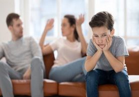 Frustrated little school boy feeling depressed while angry parents fighting at home. Worried upset small son hurt by fathers and mothers break up or divorce, children and family conflict concept.