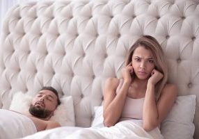 tired young woman sitting in bed with snoring husband